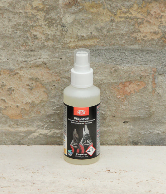 Eine kompakte Sprühflasche mit dem „FELCO Harzlöser Spray für Scheren VOC-frei Nr. 981“ von Felco steht auf einer weißen Fläche vor einer rauen, beigen Steinwand. Auf dem Etikett sind Abbildungen von Werkzeugen und Sicherheitssymbole zu sehen, die darauf hinweisen, dass das Produkt zur Reinigung von Gartengeräten (Gartenwerkzeuge reinigen) dient.