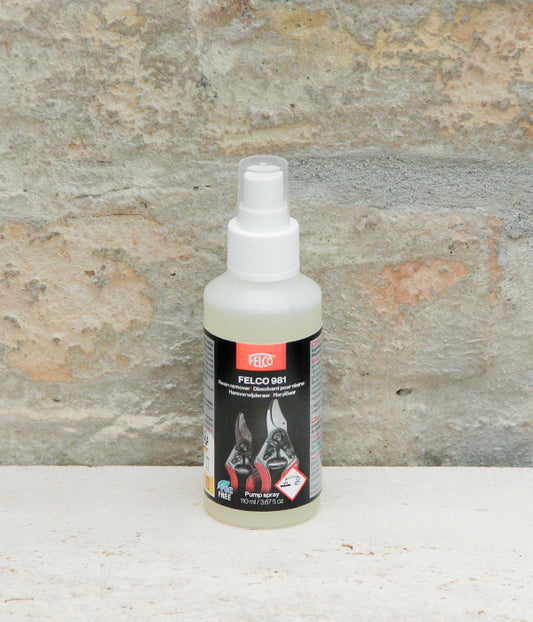 Eine Sprühflasche mit dem Produkt „FELCO Harzlöser Spray für Scheren VOC-frei Nr. 981“ von Felco, ideal zum Reinigen von Gartengeräten und zum Entfernen von Harzrückständen, mit Abbildungen von zwei Hunden auf dem Etikett vor dem Hintergrund einer Steinmauer.