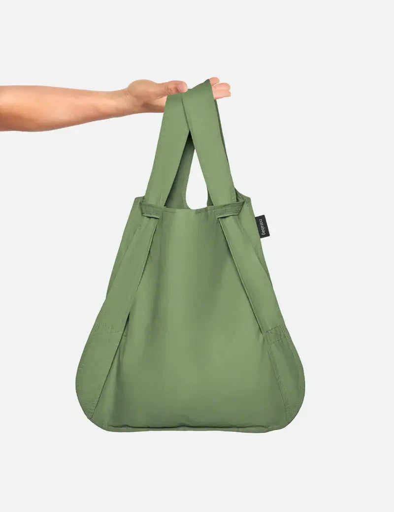 Eine Hand hält die „Notabag | Rucksack und Tasche in einem ‚Olive‘“ hoch, eine wiederverwendbare Einkaufstasche von Notabag mit zwei Tragegriffen und einer geräumigen, abgerundeten Form vor einem schlichten weißen Hintergrund. Dieses innovative Design lässt sich nahtlos von einer Tragetasche in einen Rucksack verwandeln und verbindet so Stil mit Alltagstauglichkeit.