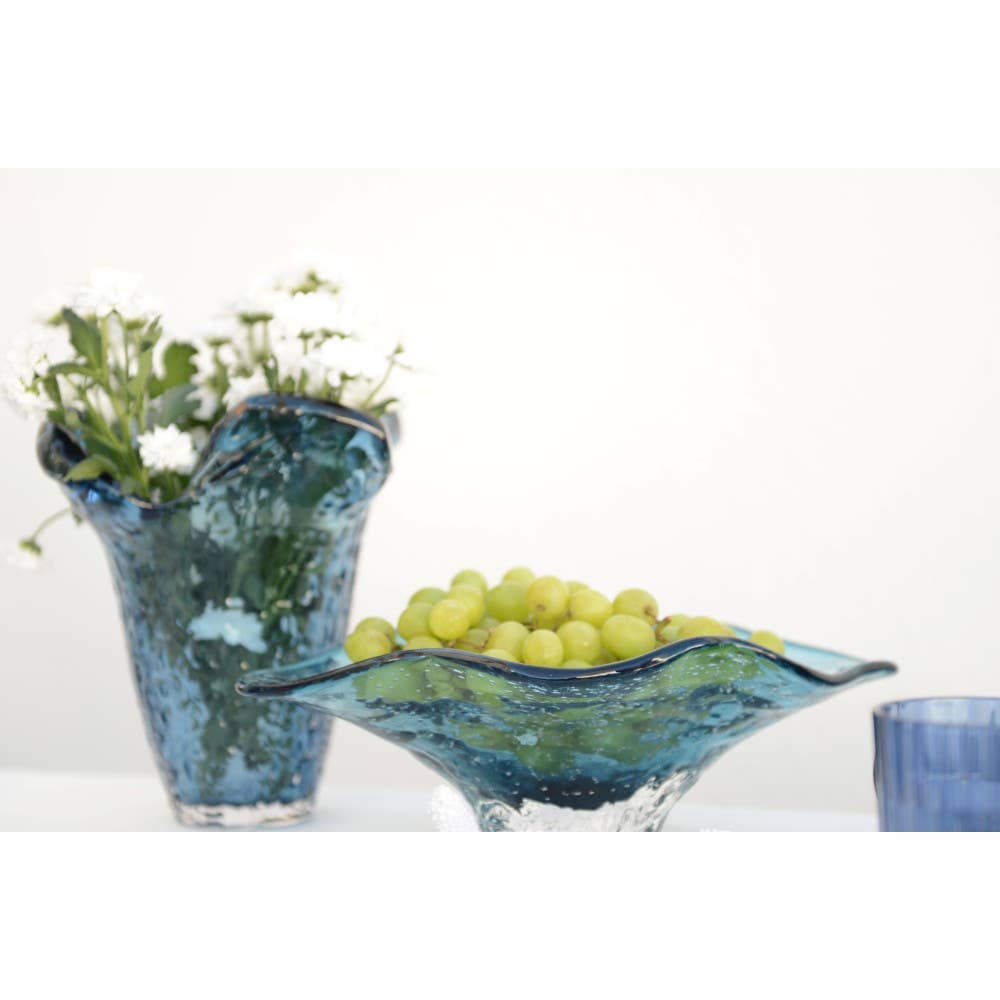 Zwei Aulica dekorative blaue Glasschale 33 x 18 x 10,5 cm - eine mit weißen Blumen und eine mit grünen Trauben - stehen auf einer weißen Fläche, neben einer weiteren dekorativen Schale und einer blauen Tasse, die rechts teilweise sichtbar ist. Marke: AULICA.