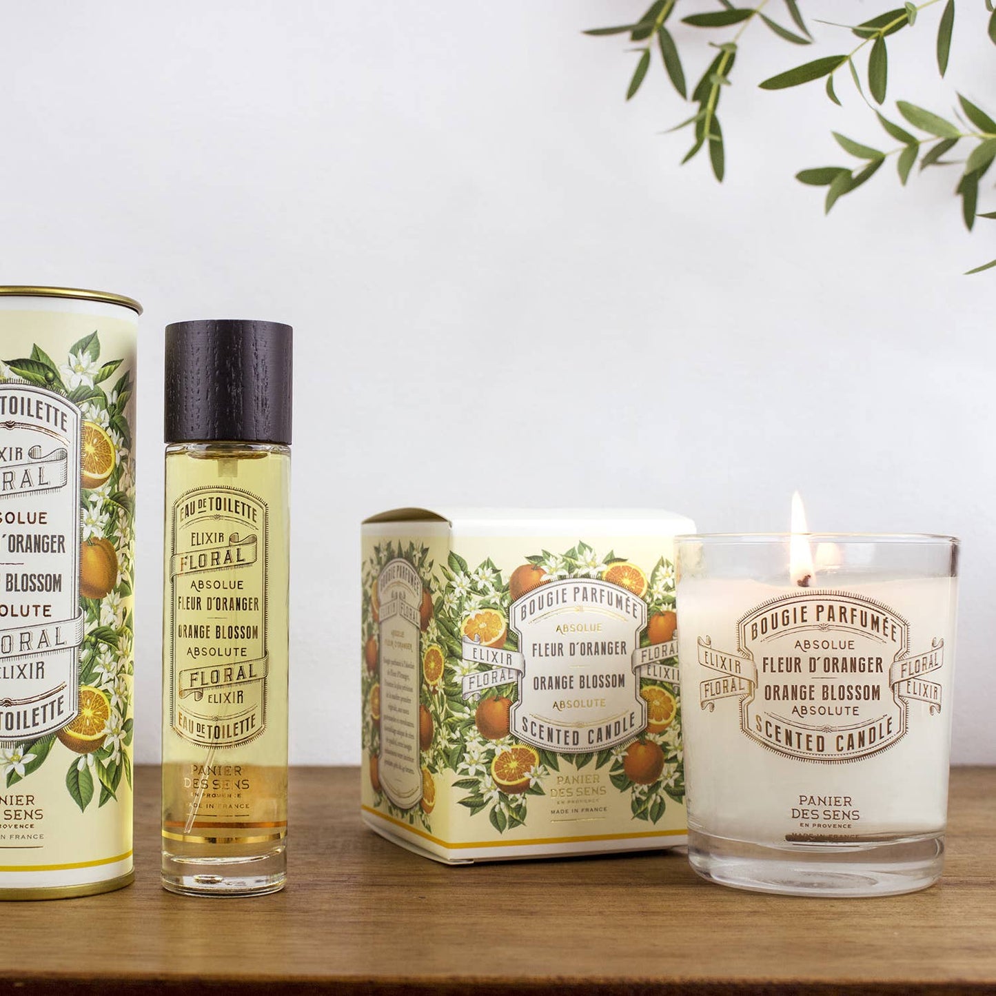 Eine Panier Des Sens Duftkerze | Orangenblüte 180 g in einer blumigen Verpackung wird auf weißem Holz präsentiert, zusammen mit einem Parfümroller; beide enthalten den veganen Duft aus Grasse.