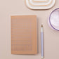 Das Tinne Mia Notizbuch A6 'Nougat Grid', ein lavendelfarbener Stift, ein rundes lilafarbenes Glas und eine ovale Keramikschale mit Goldakzenten liegen ordentlich auf hellem Beige.