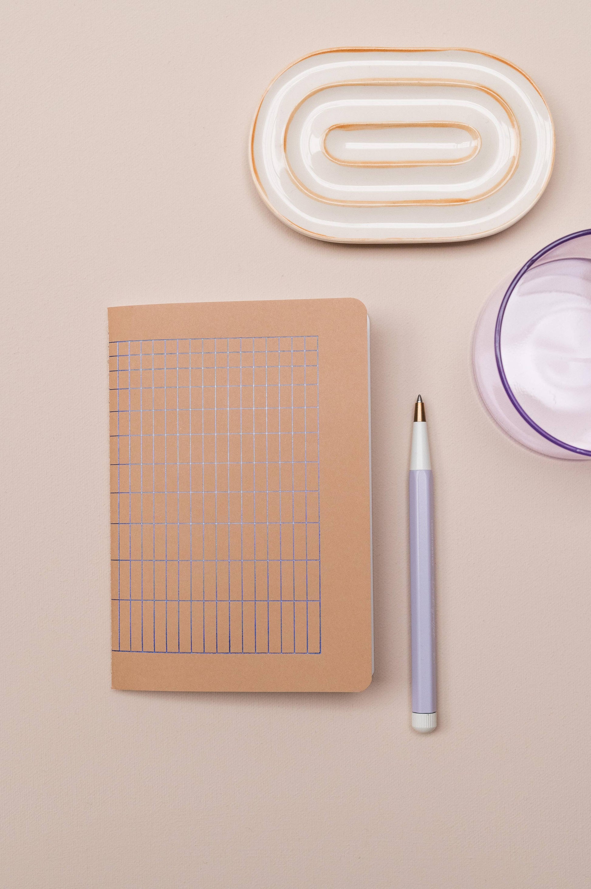 Das Tinne Mia Notizbuch A6 'Nougat Grid', ein lavendelfarbener Stift, ein rundes lilafarbenes Glas und eine ovale Keramikschale mit Goldakzenten liegen ordentlich auf hellem Beige.