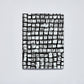 Ein Notizbuch von nuuna: das „Sassy Squares“ Notizbuch Not Ordinary L Light A5+ mit einem schwarz-weißen Einband, auf dem sich auf einem hellen, einfarbigen Hintergrund ein Raster aus unregelmäßigen, strukturierten Quadraten befindet, die wie von Hand gestempelt wirken – eine Kombination aus modernem Stil und handwerklichem Flair.
