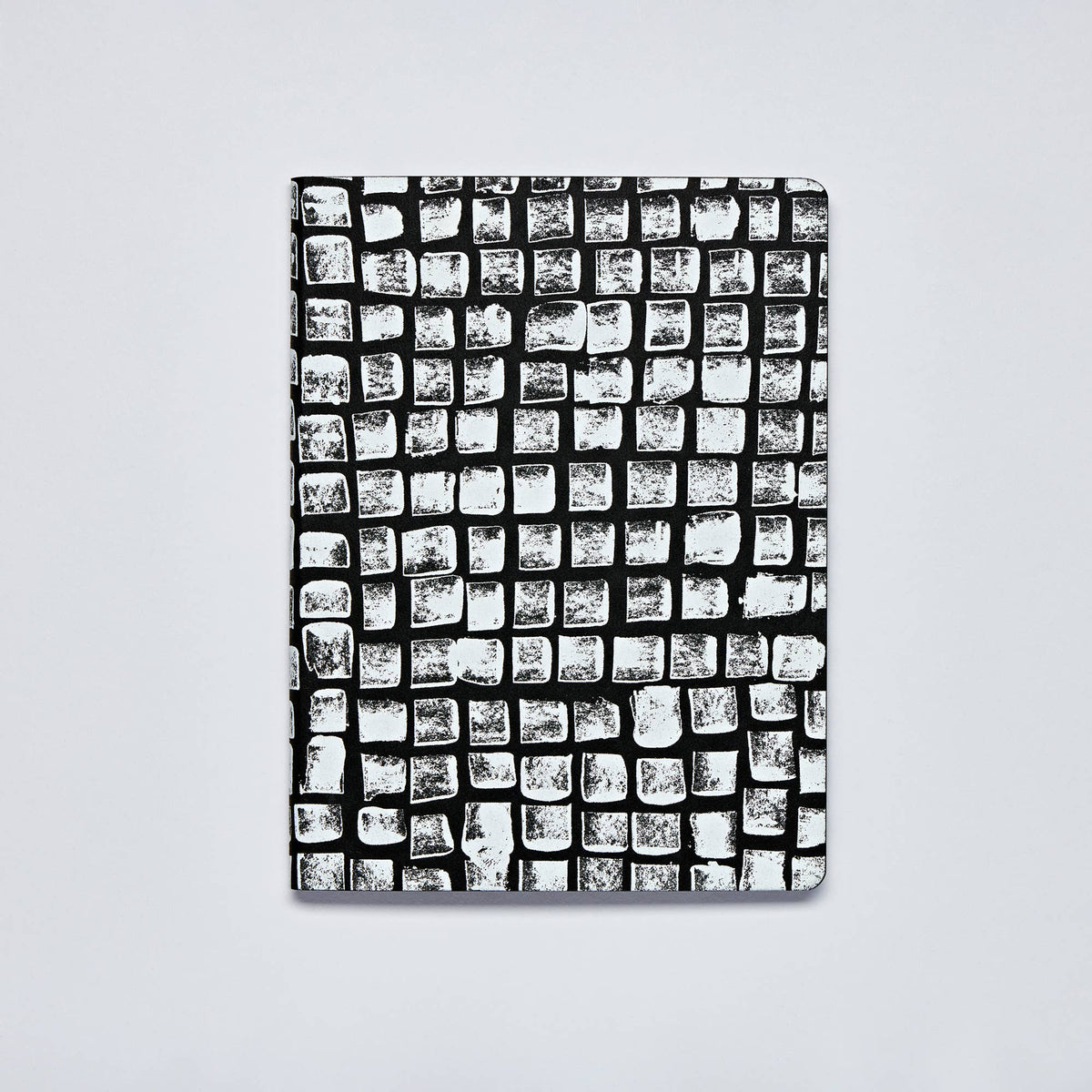 Ein Notizbuch von nuuna: das „Sassy Squares“ Notizbuch Not Ordinary L Light A5+ mit einem schwarz-weißen Einband, auf dem sich auf einem hellen, einfarbigen Hintergrund ein Raster aus unregelmäßigen, strukturierten Quadraten befindet, die wie von Hand gestempelt wirken – eine Kombination aus modernem Stil und handwerklichem Flair.
