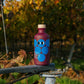 Eine lilafarbene Flasche mit blauen Kreisen, Cartoon-Augen und einer Holzkappe - Yiayia and friends Gereifter Balsamico-Essig 200 ml - steht im Freien auf Metall, während im Hintergrund grünes Gras und herbstliche Weinreben verschwimmen.