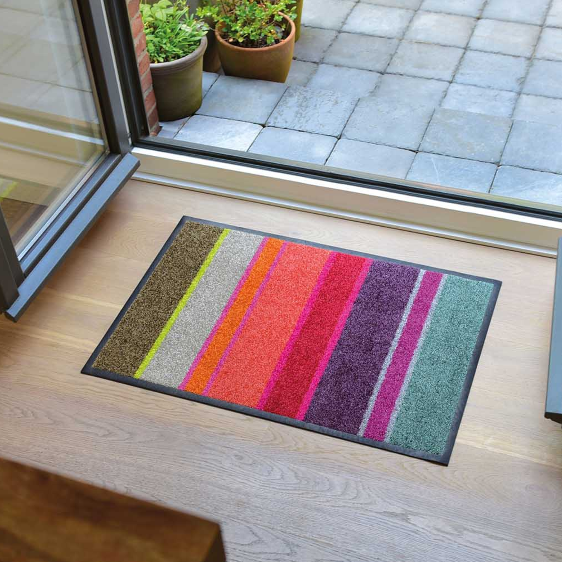 Die waschbare, rutschfeste Remember Fußmatte 'Largo' mit bunten Streifen in Grün, Gelb, Orange, Pink, Lila und Grau liegt auf Holzboden neben einer offenen Glastür; draußen sind Pflanzen und eine geflieste Terrasse sichtbar.