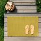 Die Fußmatte „Lida“ in Limettengrün (90 x 58 cm) von Remember ist im Innenbereich auf Holzstufen abgebildet, kombiniert mit orange-weiß gestreiften Flip-Flops und umgeben von grünem Gras. In der oberen linken Ecke der Stufen unterstreicht ein mit Brennholz gefüllter Metallkorb die gemütliche Atmosphäre, während die aus strapazierfähigen Baumwoll-Polypropylen-Mischgeweben gefertigte Fußmatte für den Innenbereich in leuchtendem Gelb hervorsticht.