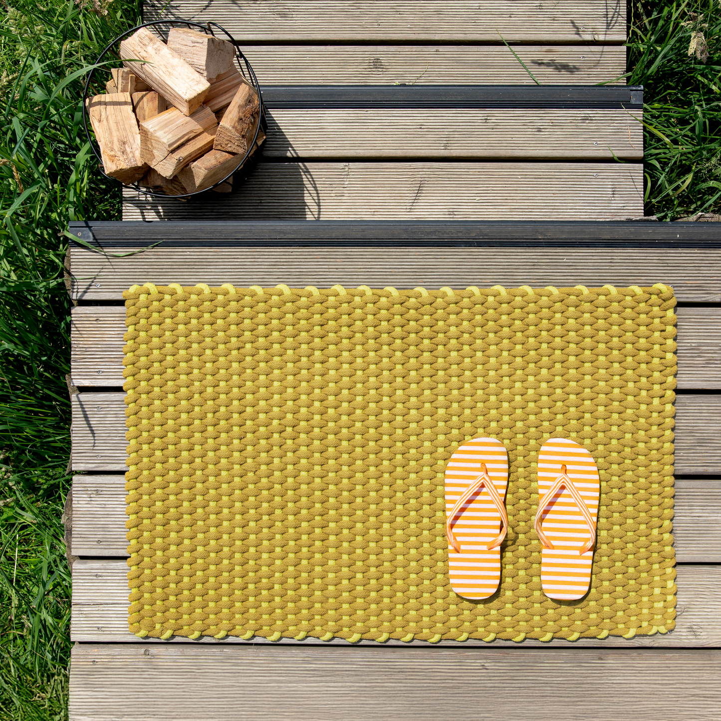 Die Fußmatte „Lida“ in Limettengrün (90 x 58 cm) von Remember ist im Innenbereich auf Holzstufen abgebildet, kombiniert mit orange-weiß gestreiften Flip-Flops und umgeben von grünem Gras. In der oberen linken Ecke der Stufen unterstreicht ein mit Brennholz gefüllter Metallkorb die gemütliche Atmosphäre, während die aus strapazierfähigen Baumwoll-Polypropylen-Mischgeweben gefertigte Fußmatte für den Innenbereich in leuchtendem Gelb hervorsticht.