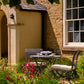 Das Harrod Horticultural Darsham Bistro-Set mit einem handgefertigten Klapptisch und zwei Stühlen steht in einem sonnenbeschienenen Garten neben einem Steinhaus, umgeben von leuchtenden Blumen und üppigem Grün, zum Tee bereit.