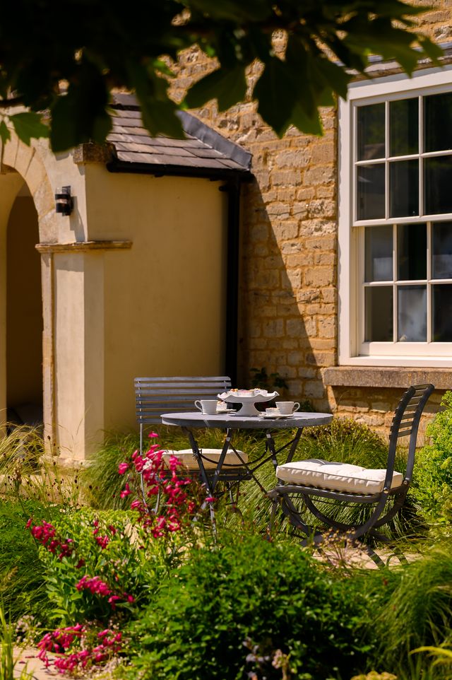 Das Harrod Horticultural Darsham Bistro-Set mit einem handgefertigten Klapptisch und zwei Stühlen steht in einem sonnenbeschienenen Garten neben einem Steinhaus, umgeben von leuchtenden Blumen und üppigem Grün, zum Tee bereit.