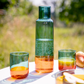 Die Remember 'Amber' Glaskaraffe aus Borosilikatglas mit grünem Schimmer und orangefarbenem Boden steht auf einem weißen Tisch neben zwei passenden Gläsern mit farbigen Getränken; im Hintergrund Natur in grünen Tönen.