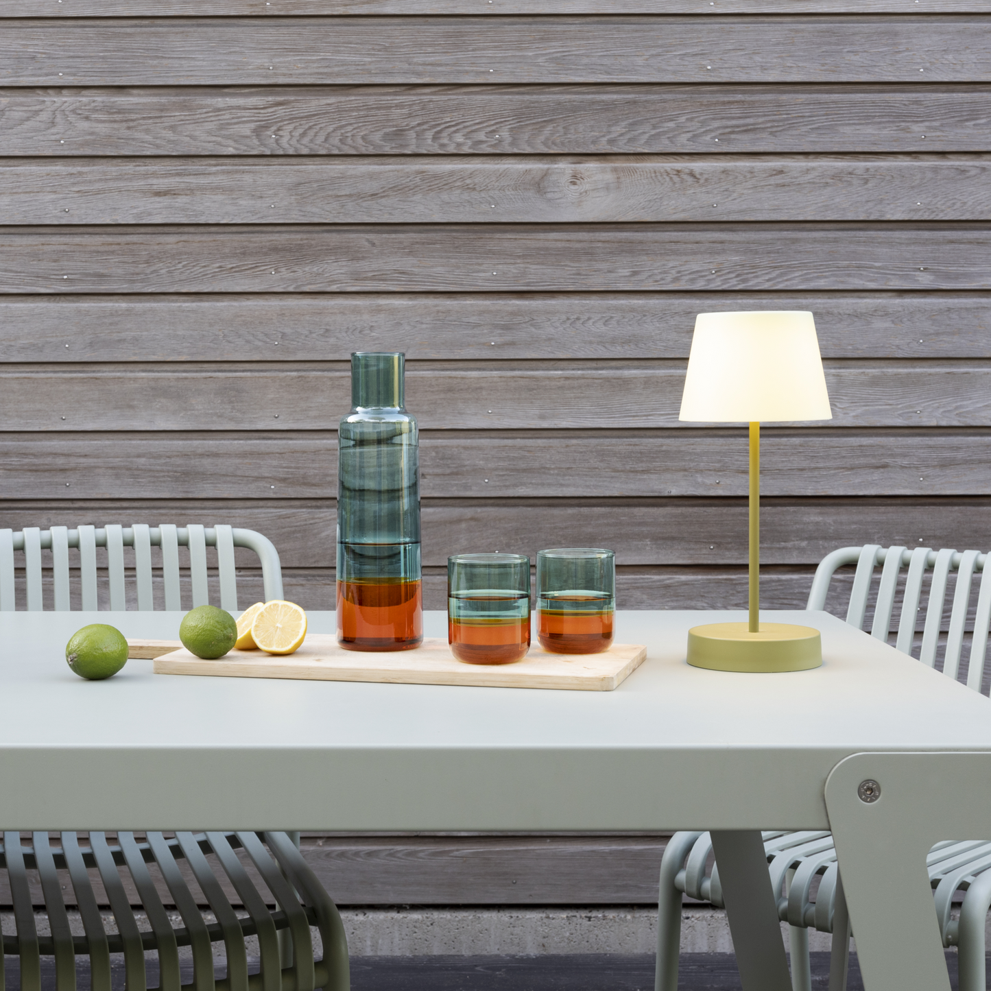 Ein moderner Outdoor-Tisch mit dem Remember 'Amber' Glaskaraffe aus Borosilikatglas, zwei Gläser Eistee, ein Schneidebrett mit Limetten und Zitronen, eine kleine Tischlampe, graue Lattenstühle und eine Holzwand schaffen eine einladende Szene.