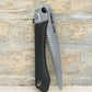 Eine Niwaki | „GR 210 Folding Saw“ Klappsäge mit schwarzem Griff und teilweise freiliegendem silbernem Sägeblatt lehnt an einer rauen, beigen Backsteinmauer und unterstreicht damit das robuste Design dieser professionellen Handsäge von Niwaki.