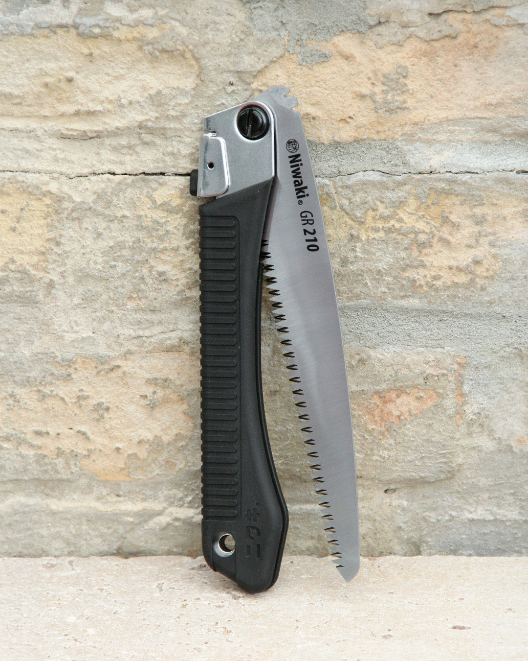 Eine Niwaki | „GR 210 Folding Saw“ Klappsäge mit schwarzem Griff und teilweise freiliegendem silbernem Sägeblatt lehnt an einer rauen, beigen Backsteinmauer und unterstreicht damit das robuste Design dieser professionellen Handsäge von Niwaki.