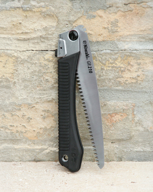 Eine Niwaki | „GR 210 Folding Saw“ Klappsäge mit schwarzem Griff und teilweise freiliegendem silbernem Sägeblatt lehnt an einer rauen, beigen Backsteinmauer und unterstreicht damit das robuste Design dieser professionellen Handsäge von Niwaki.