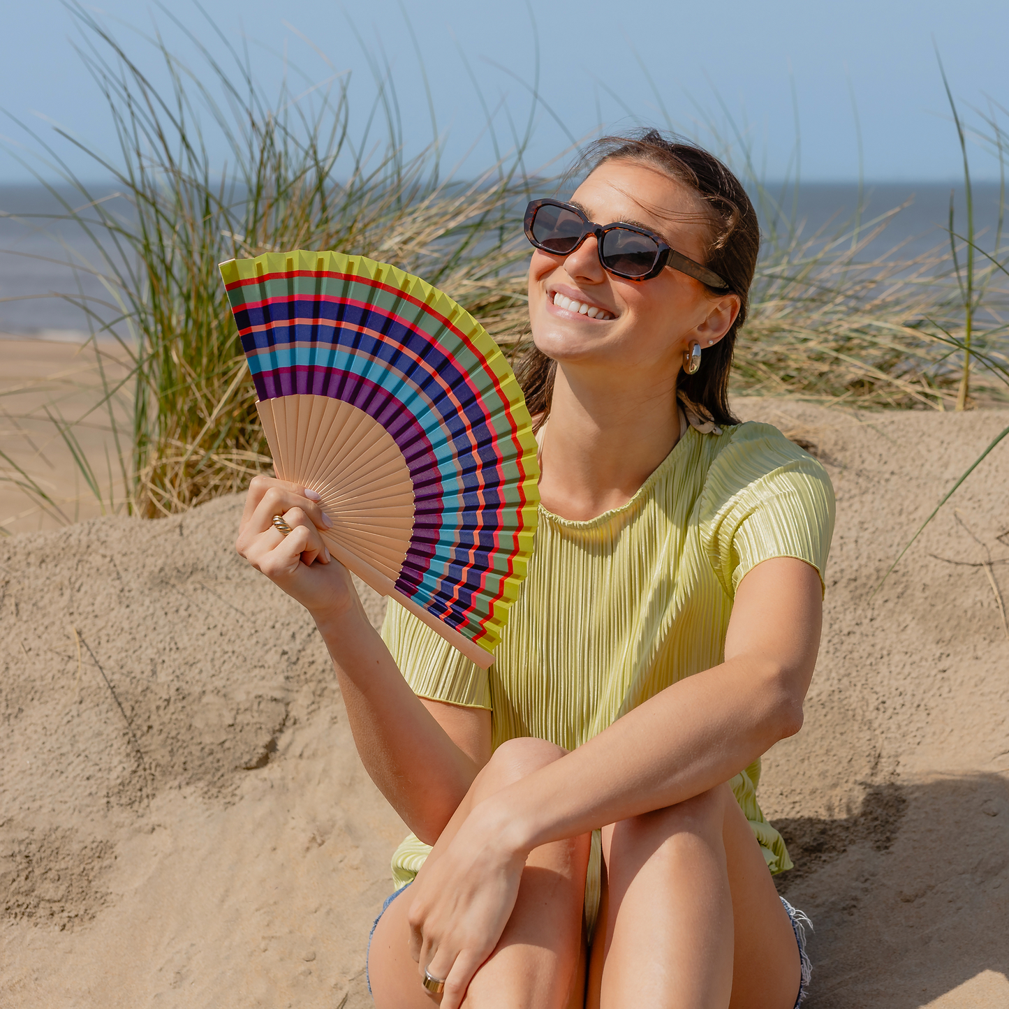 Eine lächelnde Frau mit Sonnenbrille sitzt auf einer Sanddüne am Meer und hält das farbenfrohe Remember | Handfächer 'Costa' von Remember als schickes Fächer Accessoire in der Hand, mit hohem Gras und Wellen im Hintergrund.