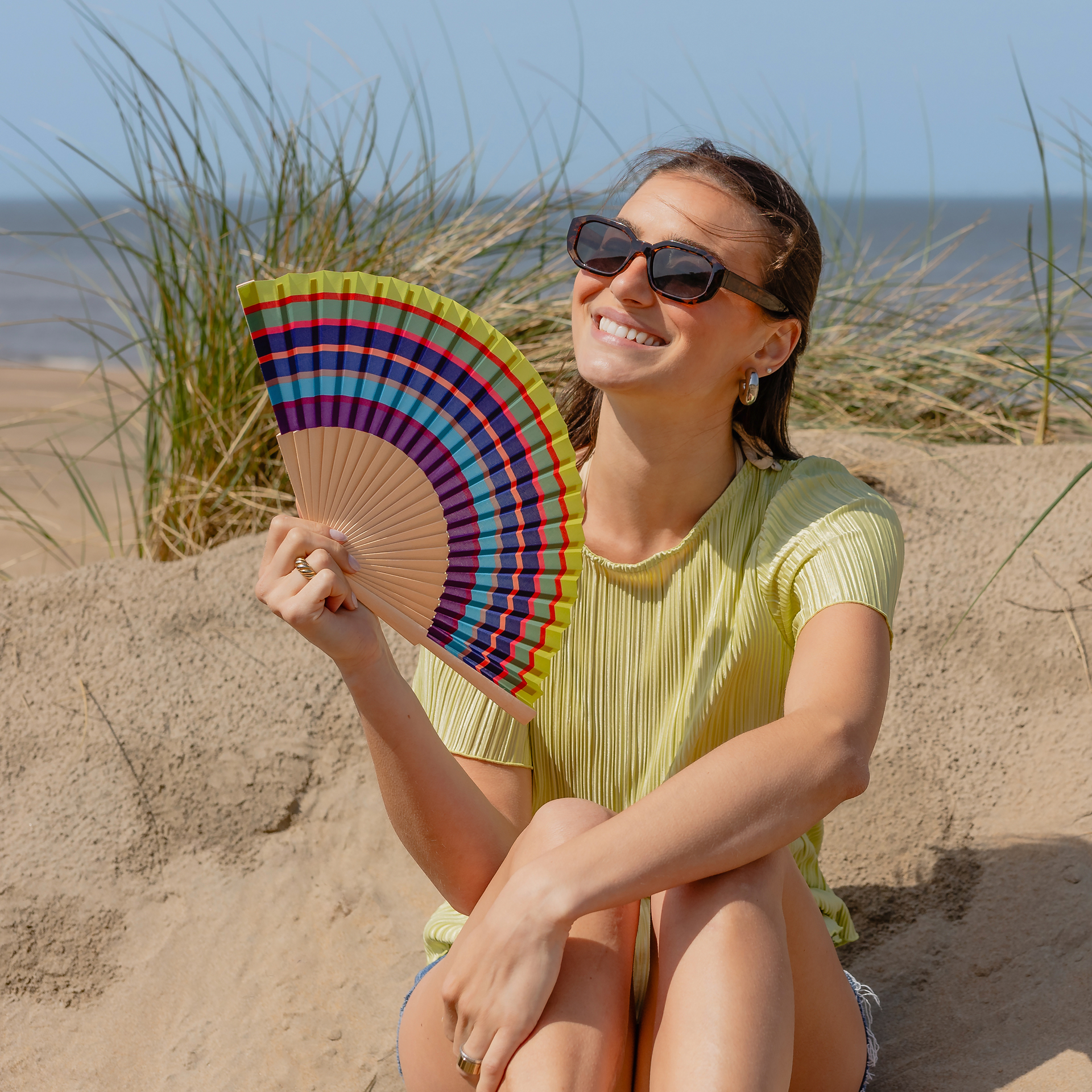 Eine lächelnde Frau mit Sonnenbrille sitzt auf einer Sanddüne am Meer und hält das farbenfrohe Remember | Handfächer 'Costa' von Remember als schickes Fächer Accessoire in der Hand, mit hohem Gras und Wellen im Hintergrund.