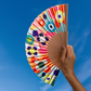 Eine Hand hält den Remember-Handfächer „Sushi“ von Remember, der sich durch ein lebhaftes, abstraktes Muster aus Kreisen und Formen vor einem strahlend blauen Himmel auszeichnet – ein stilvolles Accessoire und eine originelle Geschenkidee für jeden Anlass.