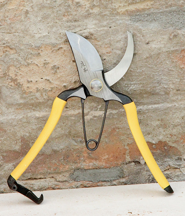Eine Niwaki-Gartenschere vom Typ „Higurashi GR“ mit gelben Griffen steht vor einer rauen Steinmauer.