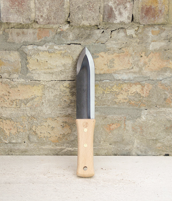 Ein unverzichtbares Hilfsmittel für die Gartenarbeit: Das Niwaki | „Hori Hori“ – ein multifunktionales Pflanzmesser mit Schutzhülle, das sich durch einen Holzgriff und eine breite, spitze Klinge auszeichnet – steht aufrecht auf einer weißen Fläche vor einer strukturierten, hellbraunen Ziegelwand.
