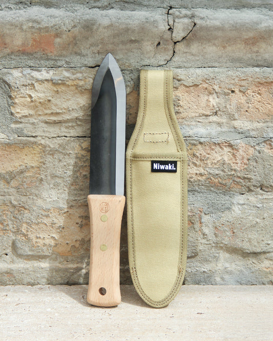 Ein Niwaki | Das multifunktionale Pflanzmesser „Hori Hori“ mit Schutzhülle steht mit seinem Holzgriff aufrecht neben der beigen Schutzhülle, beide vor einer rauen Ziegelwand platziert – ein unverzichtbares Werkzeug von Niwaki für präzise Gartenarbeit.