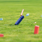 Blaue und rote rechteckige Holzklötze von Remember | KUBB Wikingerschach Gartenspiel aus Holz liegen verstreut auf grünem Gras, wobei ein blauer Klotz in der Luft liegt, als wäre er gerade getroffen worden. Der Hintergrund ist ein unscharfes Grasfeld. Marke: Remember.