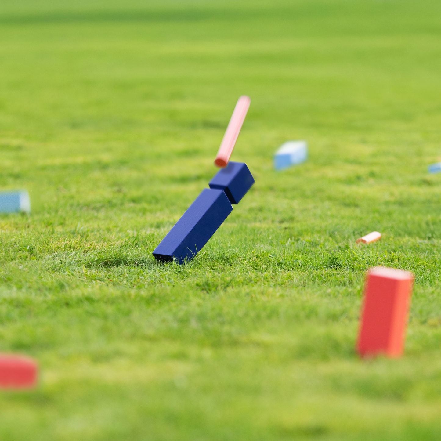Blaue und rote rechteckige Holzklötze von Remember | KUBB Wikingerschach Gartenspiel aus Holz liegen verstreut auf grünem Gras, wobei ein blauer Klotz in der Luft liegt, als wäre er gerade getroffen worden. Der Hintergrund ist ein unscharfes Grasfeld. Marke: Remember.
