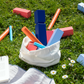 Remember | KUBB Wikingerschach Gartenspiel aus Holz von Remember zeigt farbenfrohe Holzklötze und Zylinder, die auf grünem Gras verstreut sind. Einige Teile befinden sich in einer weißen Stofftasche neben einem Buch und Gänseblümchen auf einer Picknickdecke im Sonnenschein.