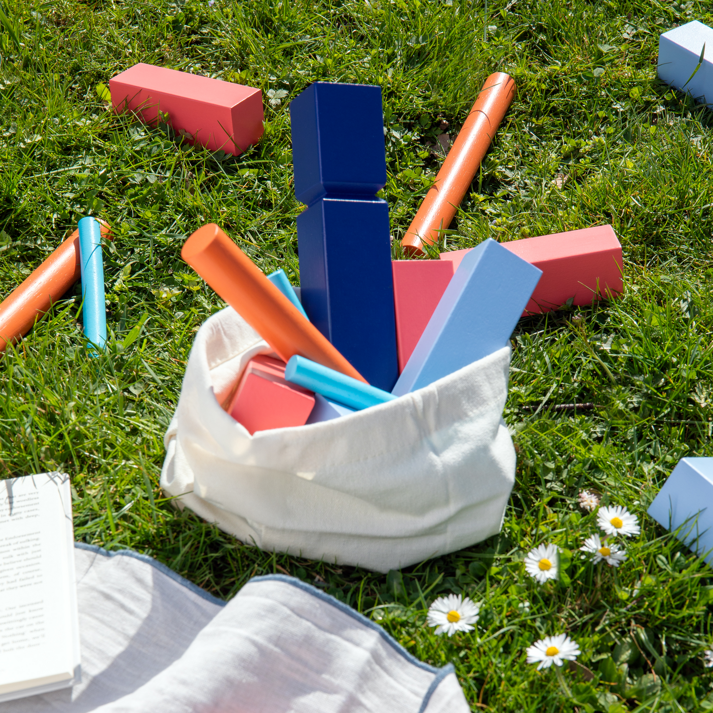 Remember | KUBB Wikingerschach Gartenspiel aus Holz von Remember zeigt farbenfrohe Holzklötze und Zylinder, die auf grünem Gras verstreut sind. Einige Teile befinden sich in einer weißen Stofftasche neben einem Buch und Gänseblümchen auf einer Picknickdecke im Sonnenschein.