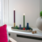 Ein moderner Kerzenhalter mit den farbenfrohen „Remember | LED-Stabkerzen Ruka 2er-Set“ von Remember, Grünpflanzen, Tannenzapfen und Walnüssen steht auf einer weißen Fensterbank neben einer runden Vase und einem Stuhl mit einem rosa Kissen.