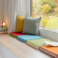 Ein gemütlicher Fensterplatz mit farbenfrohen Remember Sitzkissen 'Peanut' 40 x 40 x 5 cm, ein Stapel Bücher, eine kleine Lampe und ein aufgeschlagenes Buch mit Blick auf einen leuchtenden Garten - diese bequemen Remember Kissen machen die einladende Ecke komplett.