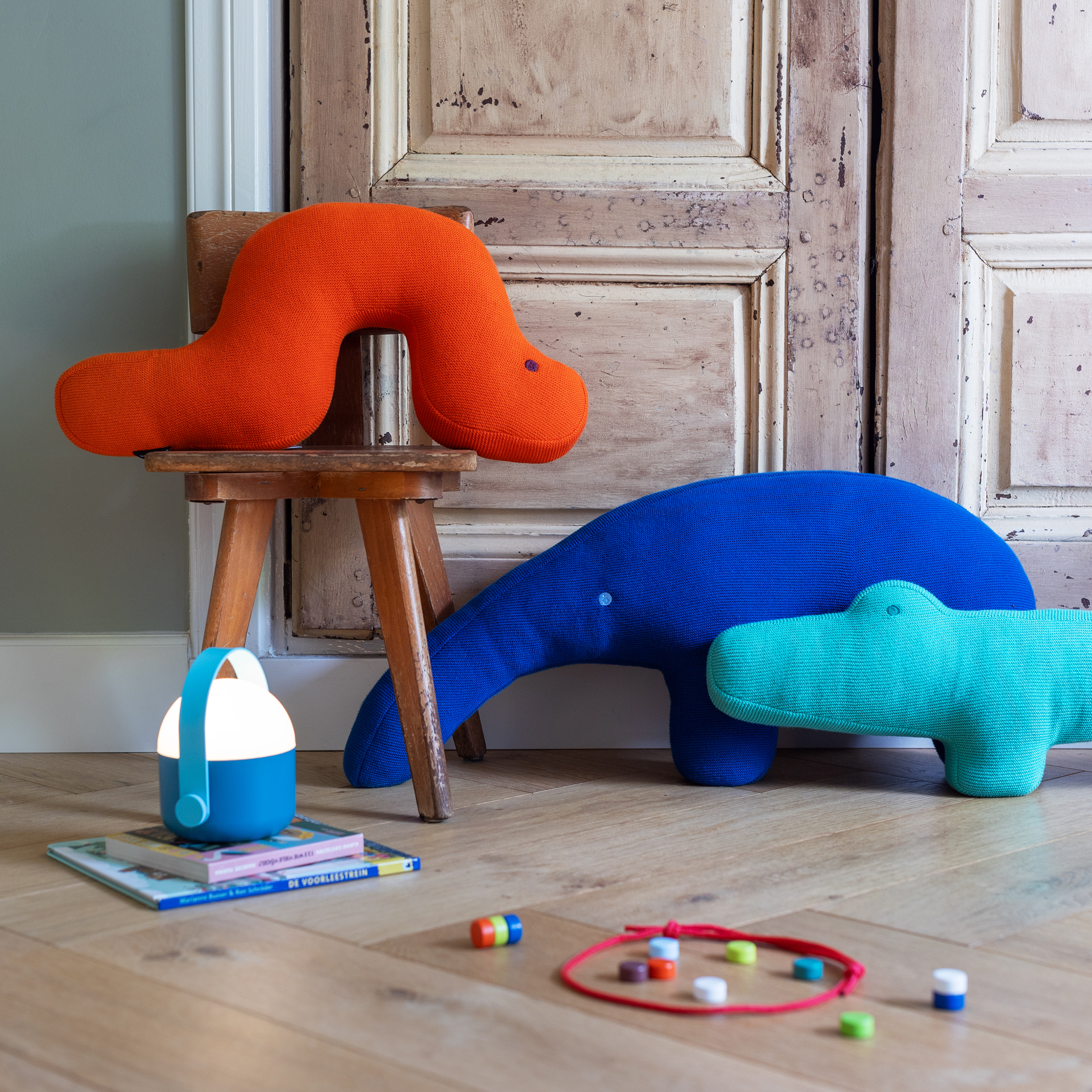 Ein farbenfrohes Kinderzimmer mit Holzboden und -tür; auf einem Holzstuhl liegt das Remember Kuschelkissen 'Ameisenbär Edgar', während blaue und blaugrüne Plüschtiere auf dem Boden liegen, umgeben von einer Laterne, Büchern und kleinen Spielsachen.