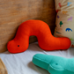 Das Remember Kuschelkissen 'Raupe Frieda' - ein leuchtend orangefarbenes, wurmförmiges Kissen mit einem einzelnen schwarzen Auge - liegt auf einem Bett neben einem türkisfarbenen Plüsch- und einem Animalprint-Kissen, ideal als liebevolle Kinderzimmer Deko.