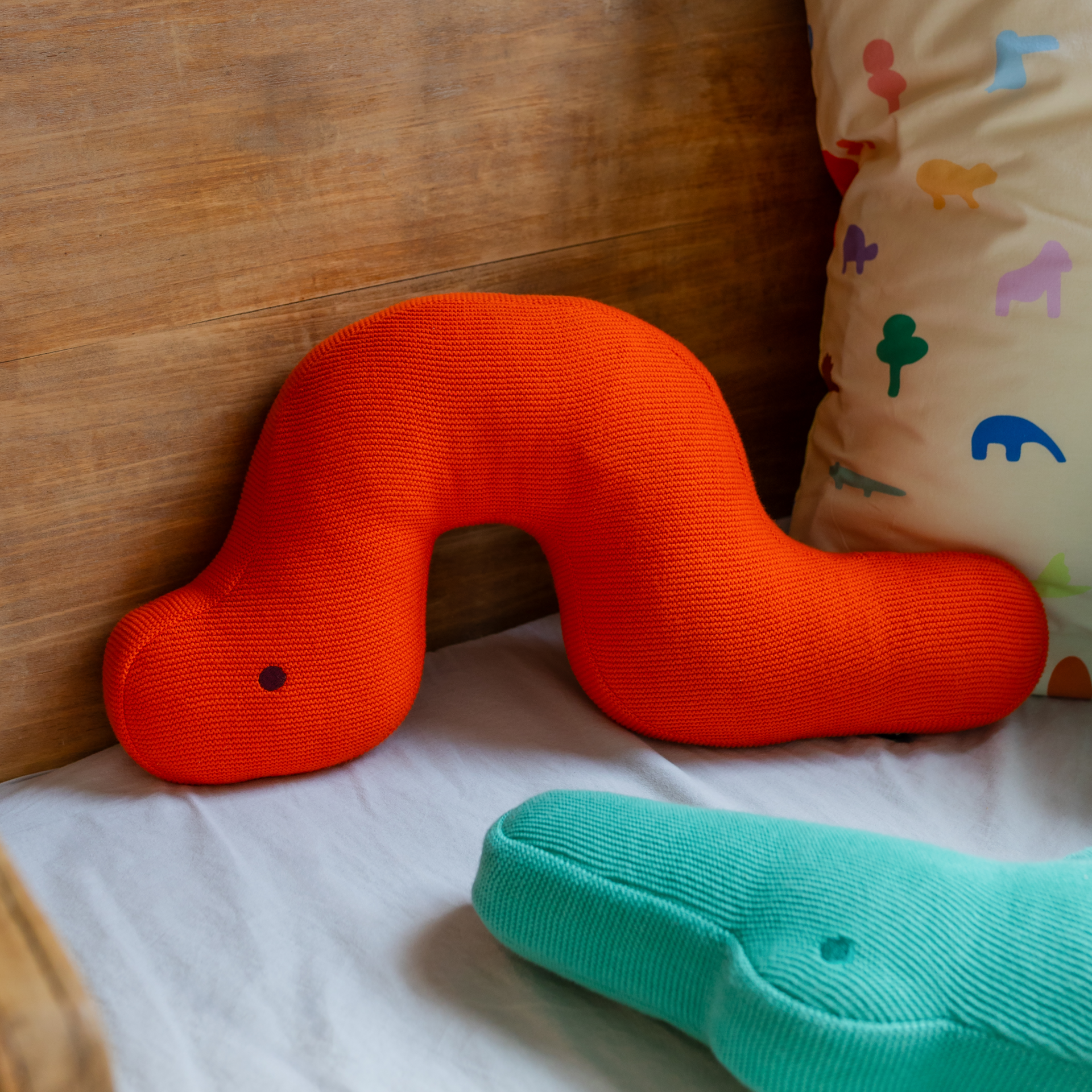 Das Remember Kuschelkissen 'Raupe Frieda' - ein leuchtend orangefarbenes, wurmförmiges Kissen mit einem einzelnen schwarzen Auge - liegt auf einem Bett neben einem türkisfarbenen Plüsch- und einem Animalprint-Kissen, ideal als liebevolle Kinderzimmer Deko.