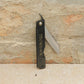 Das Niwaki | 'SK Higonokami Folding Knife' Klappmesser, mit japanischer Schrift auf dem schwarzen Griff, wird offen vor einem strukturierten Steinmauer-Hintergrund präsentiert.