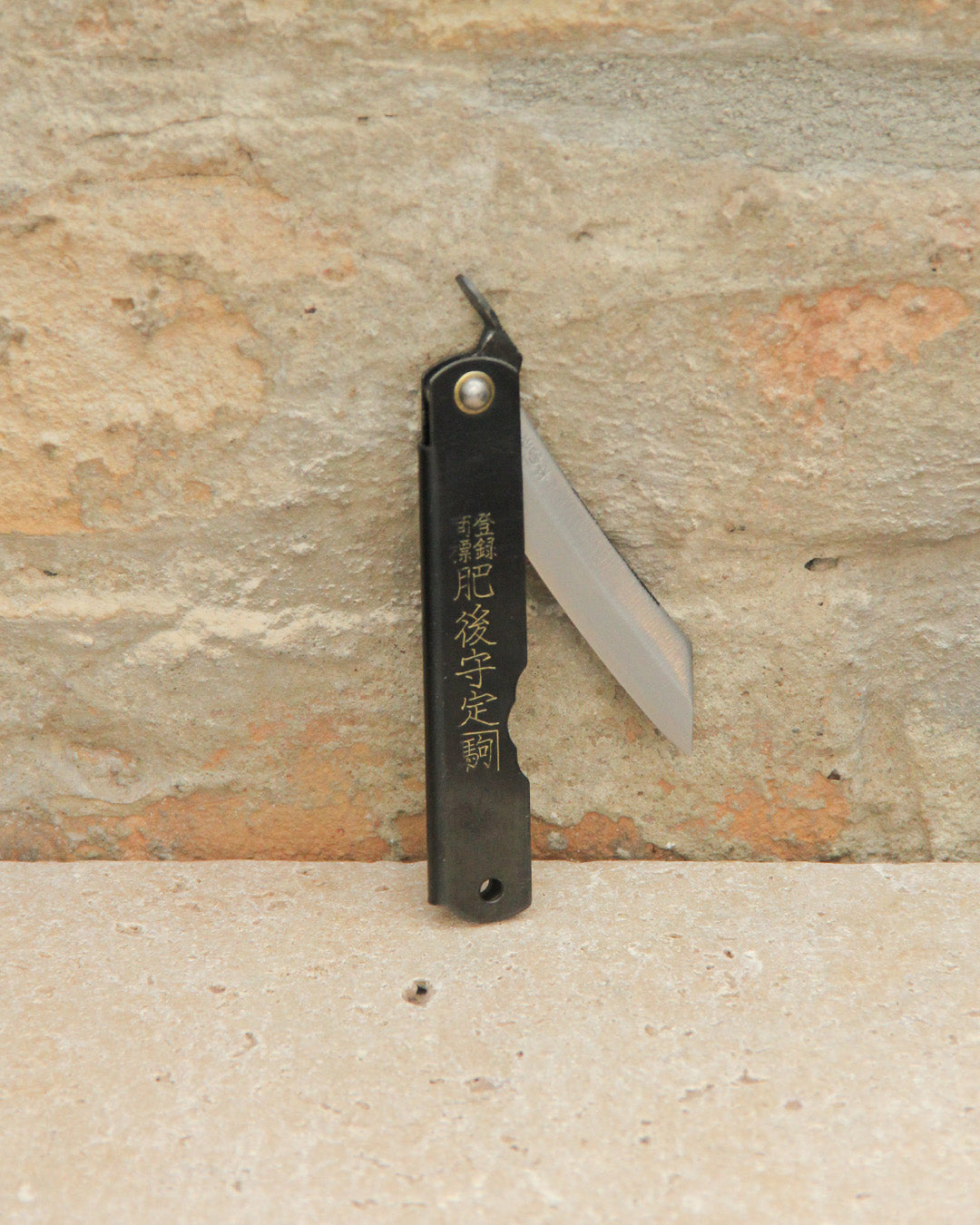 Das Niwaki | 'SK Higonokami Folding Knife' Klappmesser, mit japanischer Schrift auf dem schwarzen Griff, wird offen vor einem strukturierten Steinmauer-Hintergrund präsentiert.