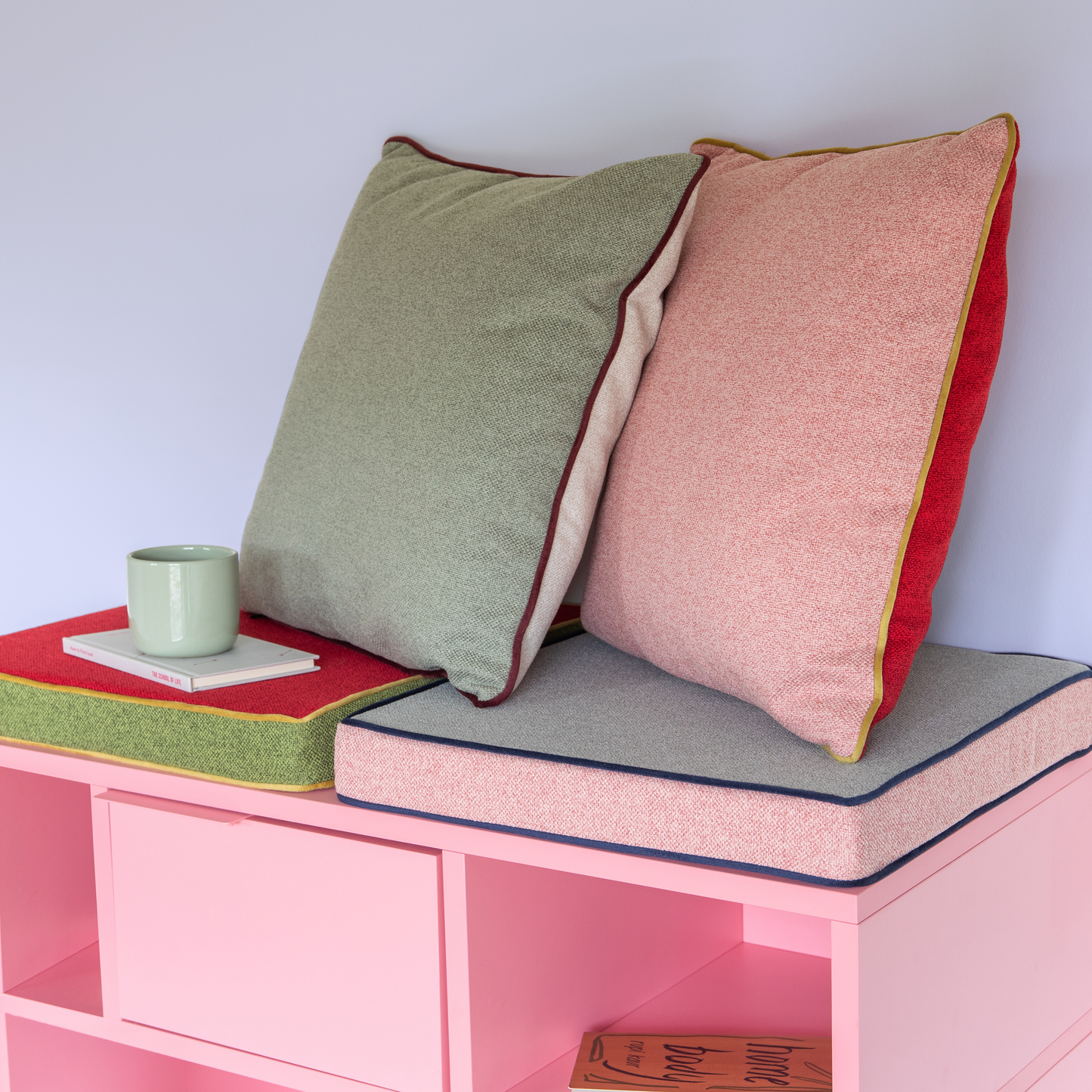 Zwei Remember Sitzkissen 'Peanut' (40x40x5cm) - eines grün, eines rosa mit farbigen Rändern - ruhen an einer Wand auf einer rosa Bank. In der Nähe steht ein grüner Becher auf einem geschlossenen Buch und ein weiteres Buch lugt aus dem Inneren der Bank hervor.
