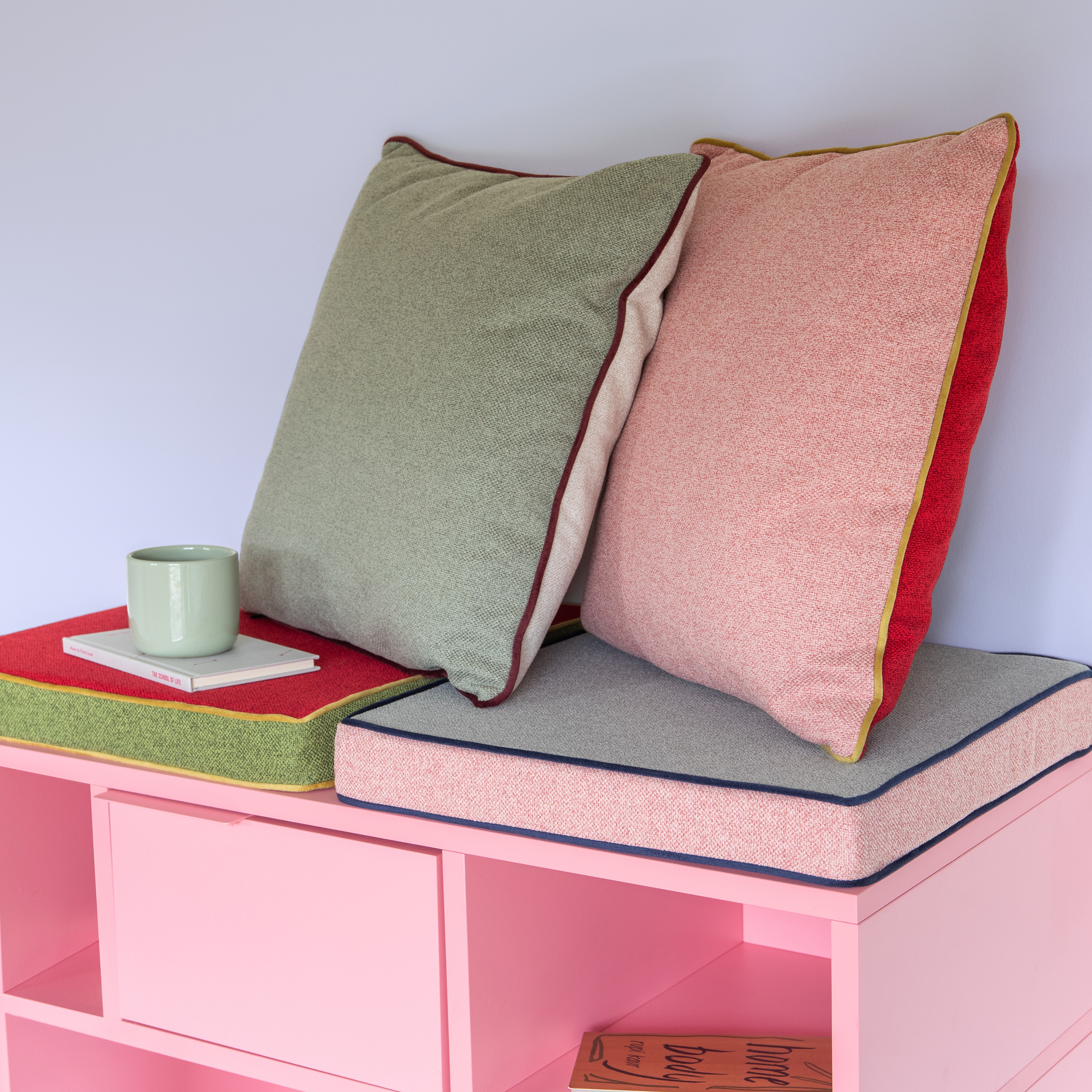 Zwei Remember Sitzkissen 'Peanut' (40x40x5cm) - eines grün, eines rosa mit farbigen Rändern - ruhen an einer Wand auf einer rosa Bank. In der Nähe steht ein grüner Becher auf einem geschlossenen Buch und ein weiteres Buch lugt aus dem Inneren der Bank hervor.