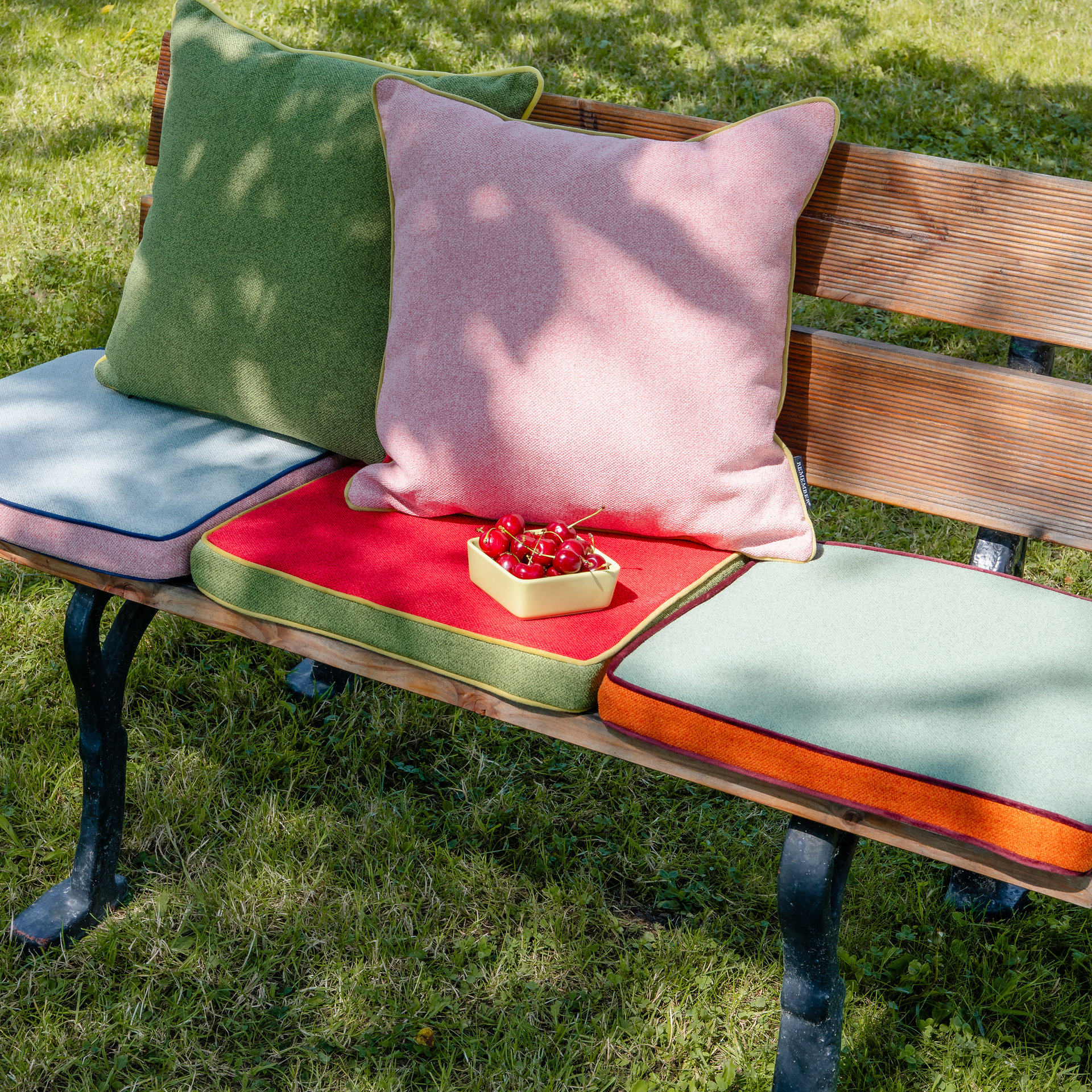 Eine Holzbank mit Metallbeinen ist mit einem Remember Sitzkissen 'Cranberry' 40x40x5 cm von Remember belegt. Eine kleine gelbe Schale, gefüllt mit Kirschen, steht auf dem roten Kissen, während die Bank auf Gras im Schatten eines Baumes ruht.