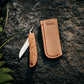 Ein Niwaki | 'Kotoh X Niwaki Knife' Taschenmesser mit Schutzhülle aus Leder liegt mit geöffnetem Holzgriff auf einem Felsen neben der tanfarbenen, Niwaki-gebrandeten Lederschutzhülle, umgeben von grünen Farnen - ideal für Gartenarbeit.