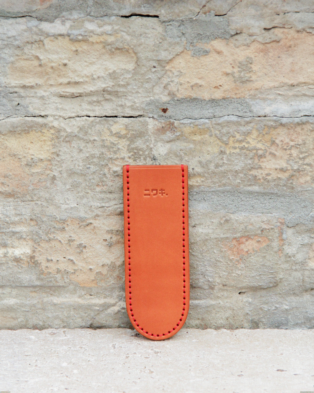 Ein hellbraunes Lederetui für Stifte oder Bleistifte mit sichtbaren Nähten und japanischen Schriftzeichen, das an die „Leather Sheath“ Lederschutzhülle für kleine Messer von Niwaki erinnert, steht aufrecht an einer strukturierten Steinwand.