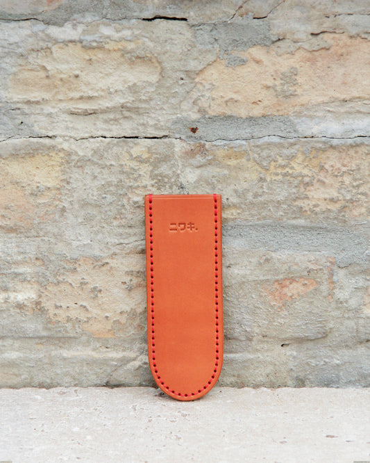 Ein hellbraunes Lederetui für Stifte oder Bleistifte mit sichtbaren Nähten und japanischen Schriftzeichen, das an die „Leather Sheath“ Lederschutzhülle für kleine Messer von Niwaki erinnert, steht aufrecht an einer strukturierten Steinwand.