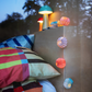 Neben einem hölzernen Kopfteil, das mit der Lichterkette „Remember Papier Lichterkette Yazz“ (150 cm) von Remember geschmückt ist, steht ein farbenfrohes Bett mit einer karierten Bettdecke und Kissen; darauf stehen zwei kleine Lampen und Papierornamente, während vor dem Fenster Bäume zu sehen sind.