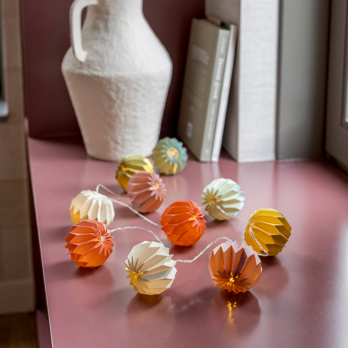 Die „Remember Papier Lichterkette Yazz“ (150 cm) von Remember zeigt orangefarbene, gelbe, weiße und grüne Papierlaternen, die auf einem rosa Untergrund in der Nähe einer weißen Vase, einiger Bücher und eines Fensters angeordnet sind. Diese farbenfrohe LED-Lichterkette erhellt jeden Raum mit ihrem fröhlichen Schein.