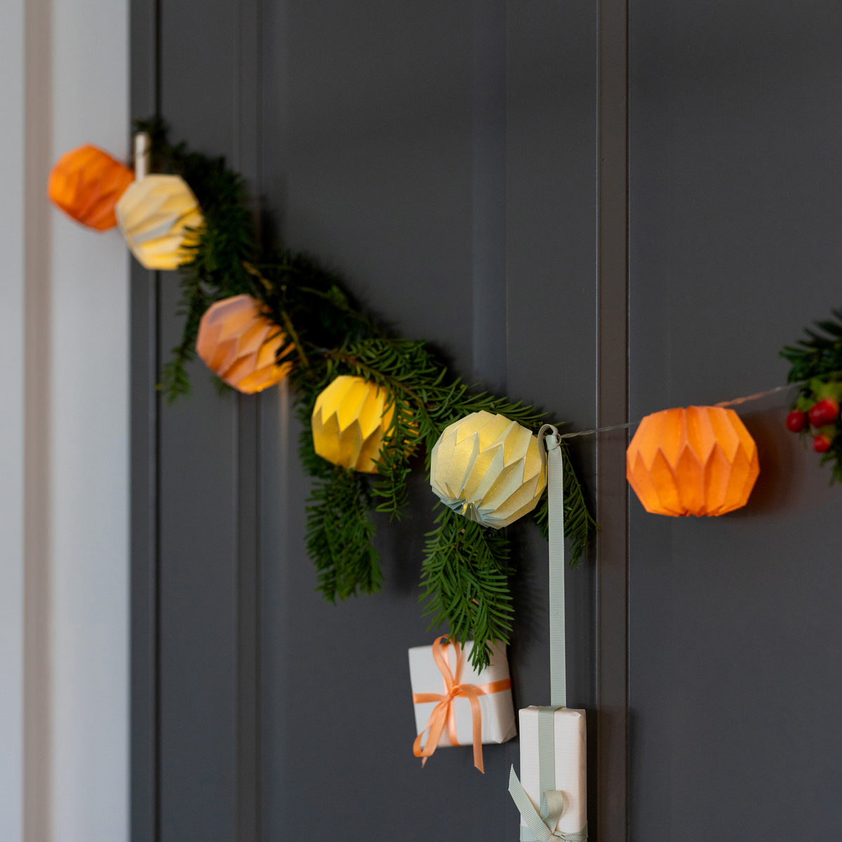 An einer dunklen Tür hängt eine Lichterkette aus orangefarbenen und gelben Papierlaternen von „Remember“, die „Papier Lichterkette Yazz“ (150 cm), geschmückt mit Grünzweigen und kleinen, verpackten Geschenken.