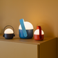 Drei moderne, tragbare „Remember | Ole“ Mini-LED-Leuchten im Dreier-Set – mit kuppelförmigen weißen Oberteilen und farbigen Sockeln in Schwarz, Blau und Rot – stehen auf einem beigen Schrank vor einer hellbraunen Wand. Jede dimmbare Leuchte von Remember verfügt über einen passenden Griff und ist akkubetrieben, was einen bequemen kabellosen Einsatz ermöglicht.
