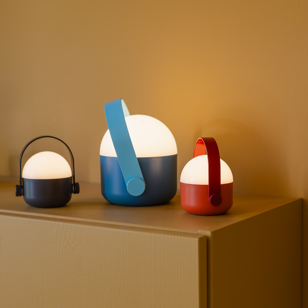 Drei moderne, tragbare „Remember | Ole“ Mini-LED-Leuchten im Dreier-Set – mit kuppelförmigen weißen Oberteilen und farbigen Sockeln in Schwarz, Blau und Rot – stehen auf einem beigen Schrank vor einer hellbraunen Wand. Jede dimmbare Leuchte von Remember verfügt über einen passenden Griff und ist akkubetrieben, was einen bequemen kabellosen Einsatz ermöglicht.