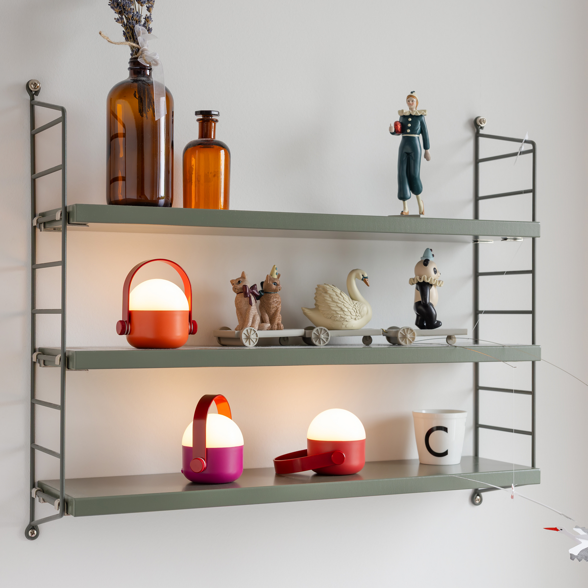 Auf drei Regalen stehen Vintage-Flaschen, Spielzeugfiguren, Schwan- und Hunde-Dekofiguren, zwei dimmbare „Remember | Ole“ Mini-LED-Leuchten in Rot von Remember – praktischerweise akkubetrieben – sowie eine Tasse mit dem Buchstaben C vor einer hellen Wand.