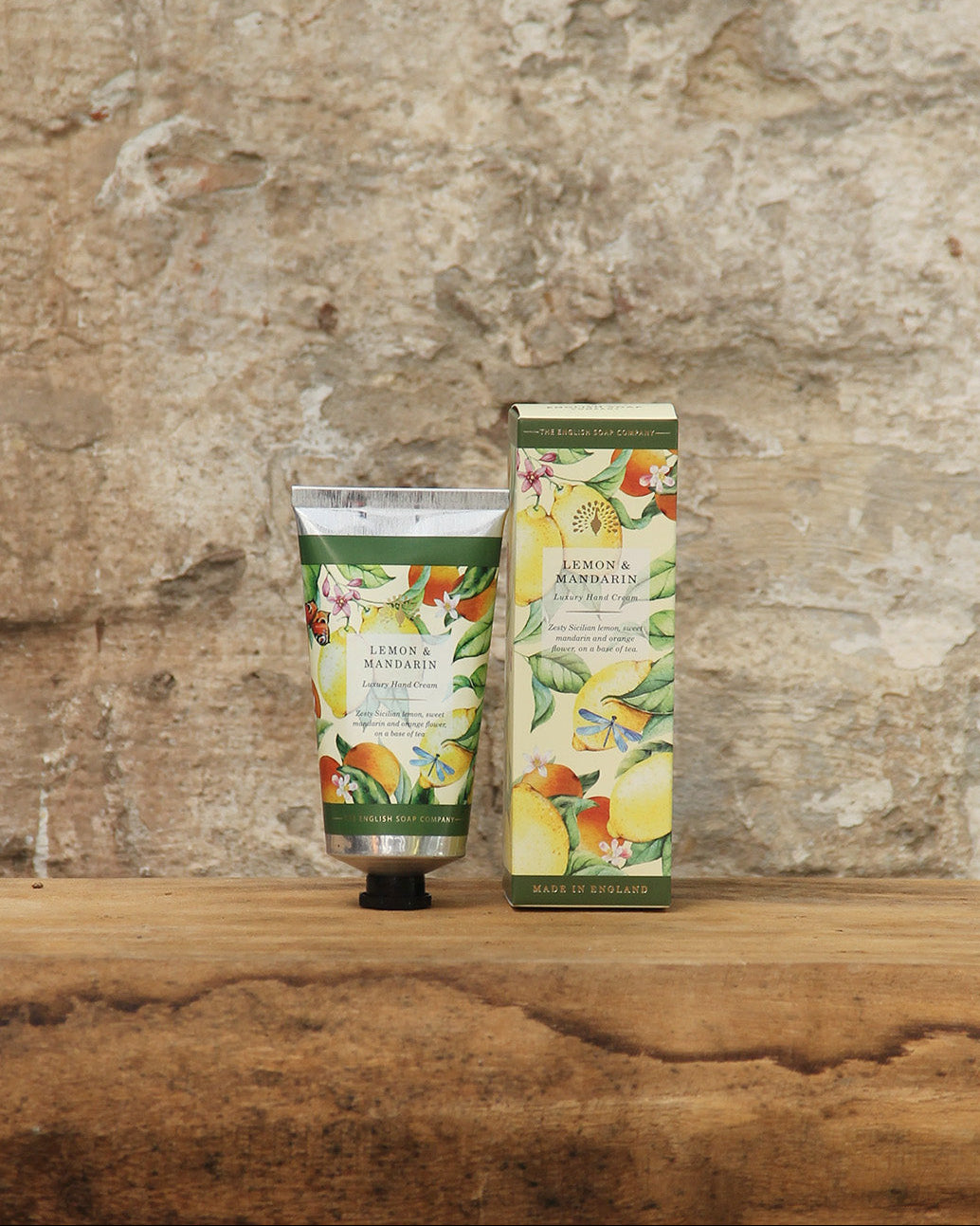 Eine Tube und eine Schachtel der ESC | 'Lemon Mandarin' Handcreme 75 ml von The English Soap Company mit lebhaften botanischen Motiven ruhen auf Holz vor einer Steinwand - eine stilvolle Kulisse für diese luxuriöse vegane Handpflege.